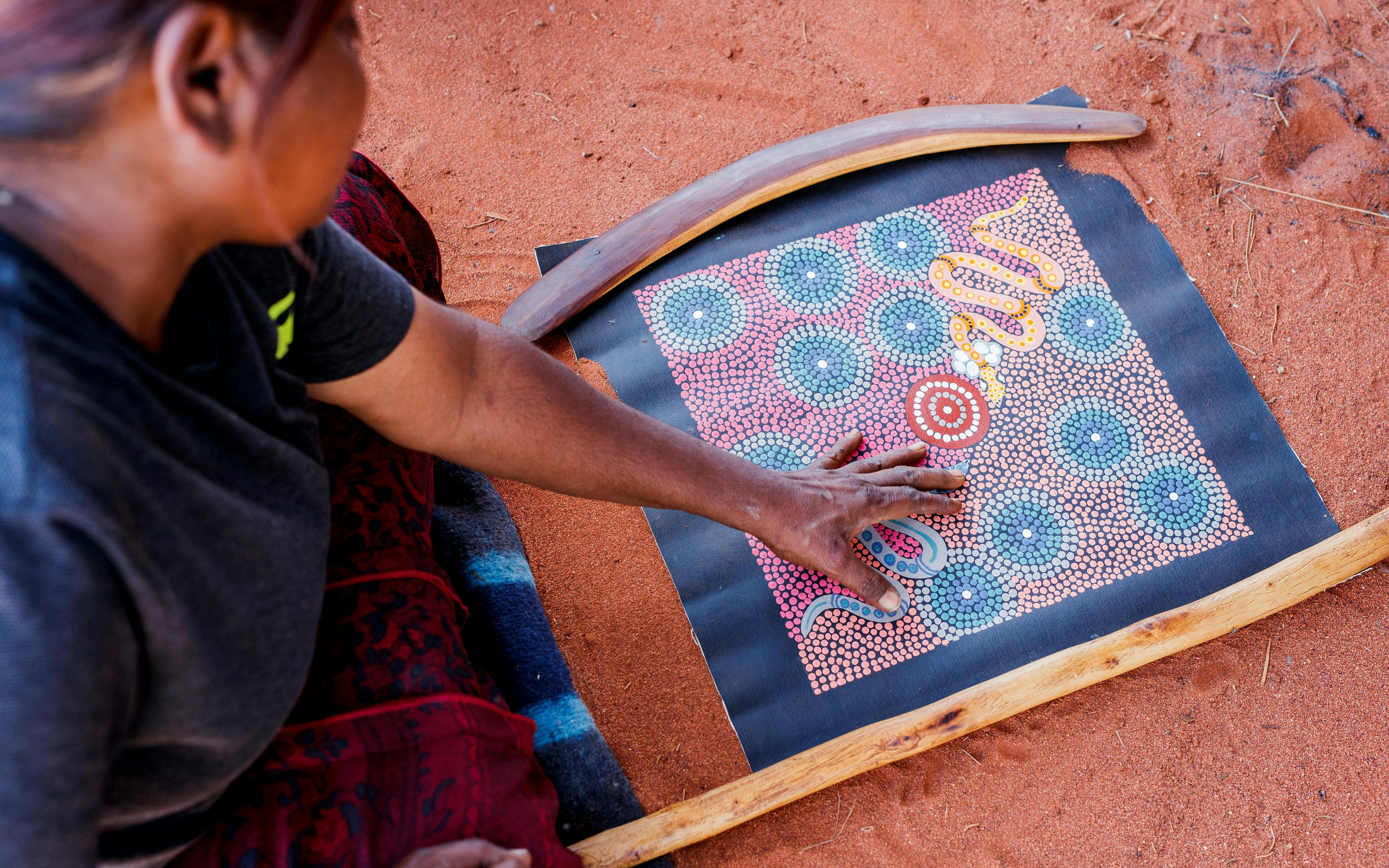 Aboriginal artist creating dot painting on red sand during Uluru tour.