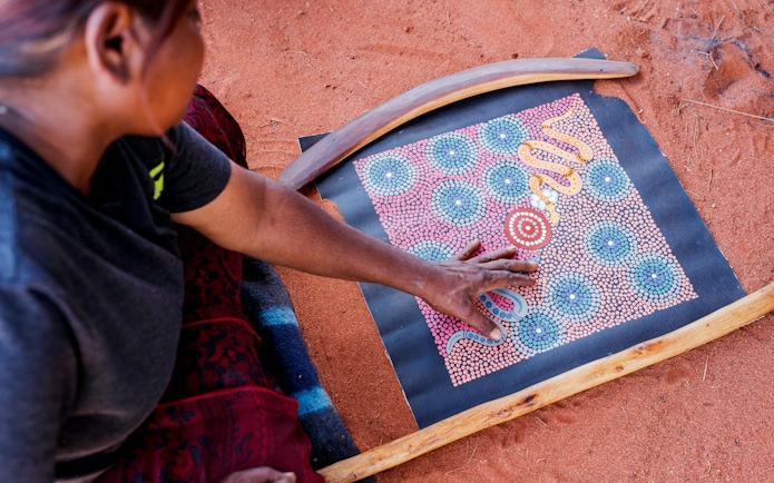Aboriginal artist creating dot painting on red sand during Uluru tour.