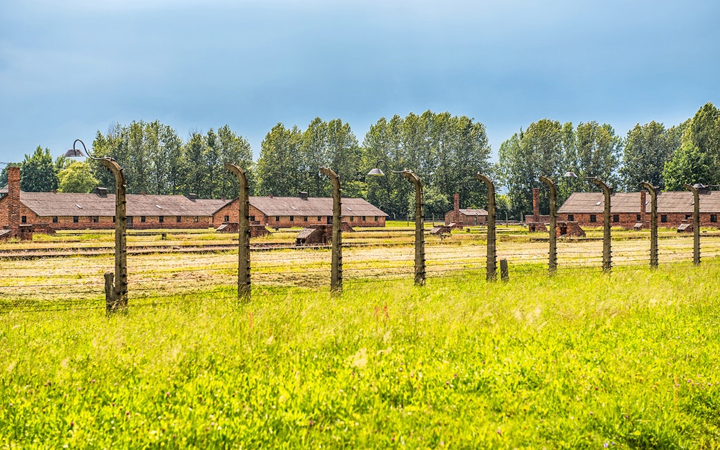 Auschwitz-Birkenau barracks and barbed wire fence under a blue sky.