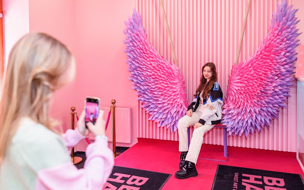 Person posing on a bench with pink and purple wings at Be Happy Museum, Krakow.
