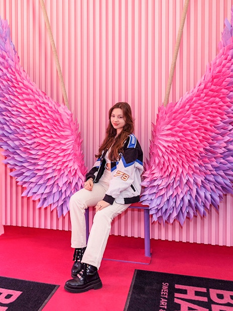 Person posing on a bench with pink and purple wings at Be Happy Museum, Krakow.