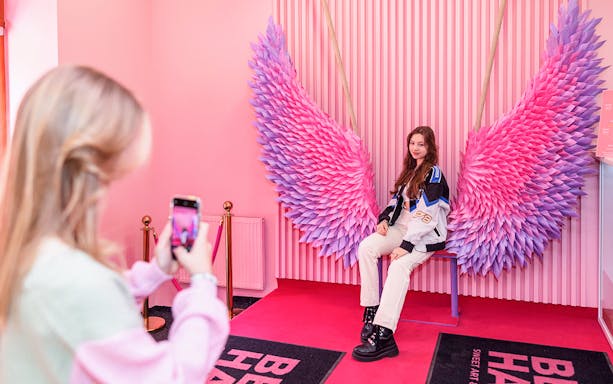 Person posing on a bench with pink and purple wings at Be Happy Museum, Krakow.