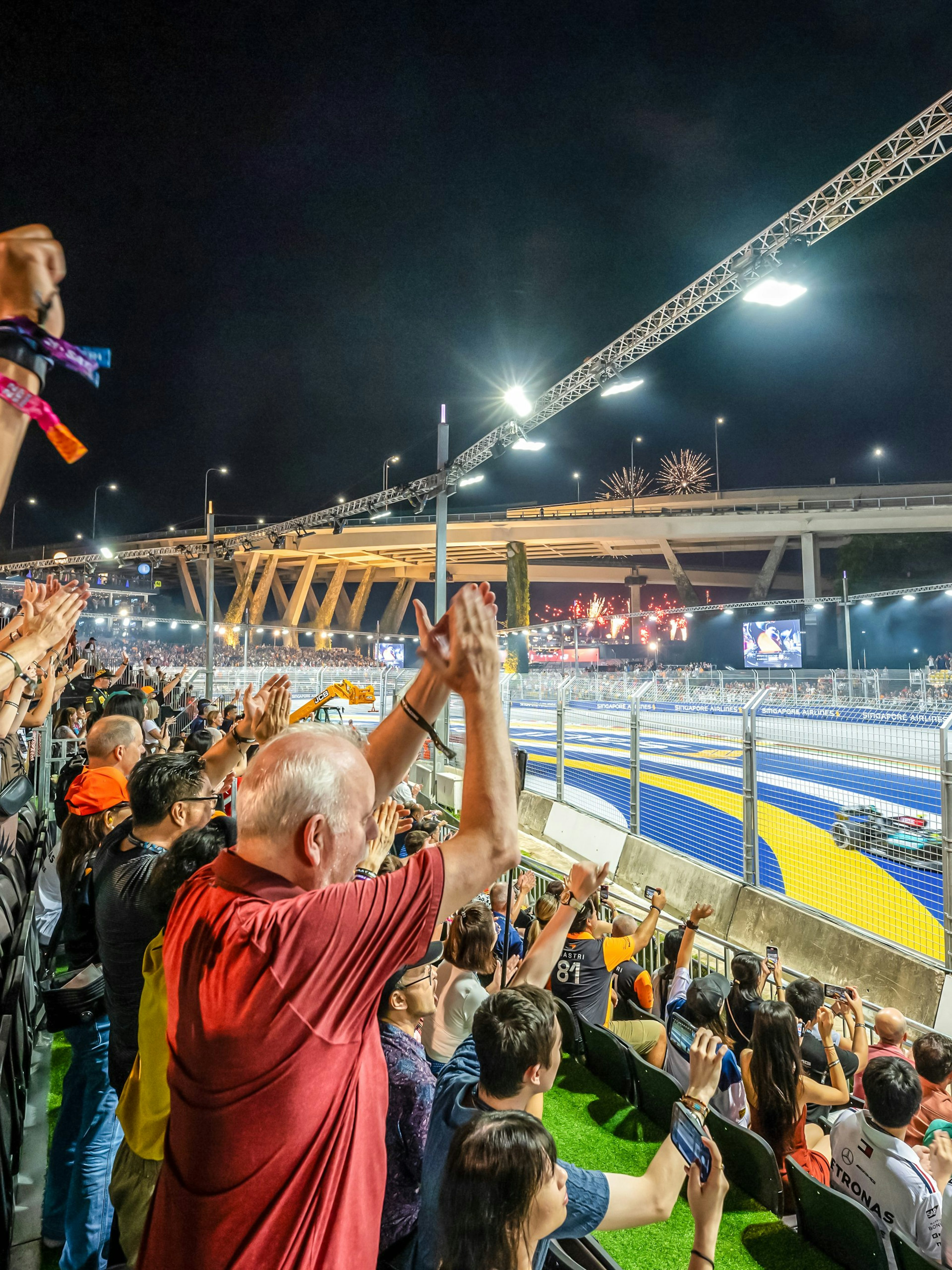Spectators cheering at a night race with fireworks and city skyline in the background.