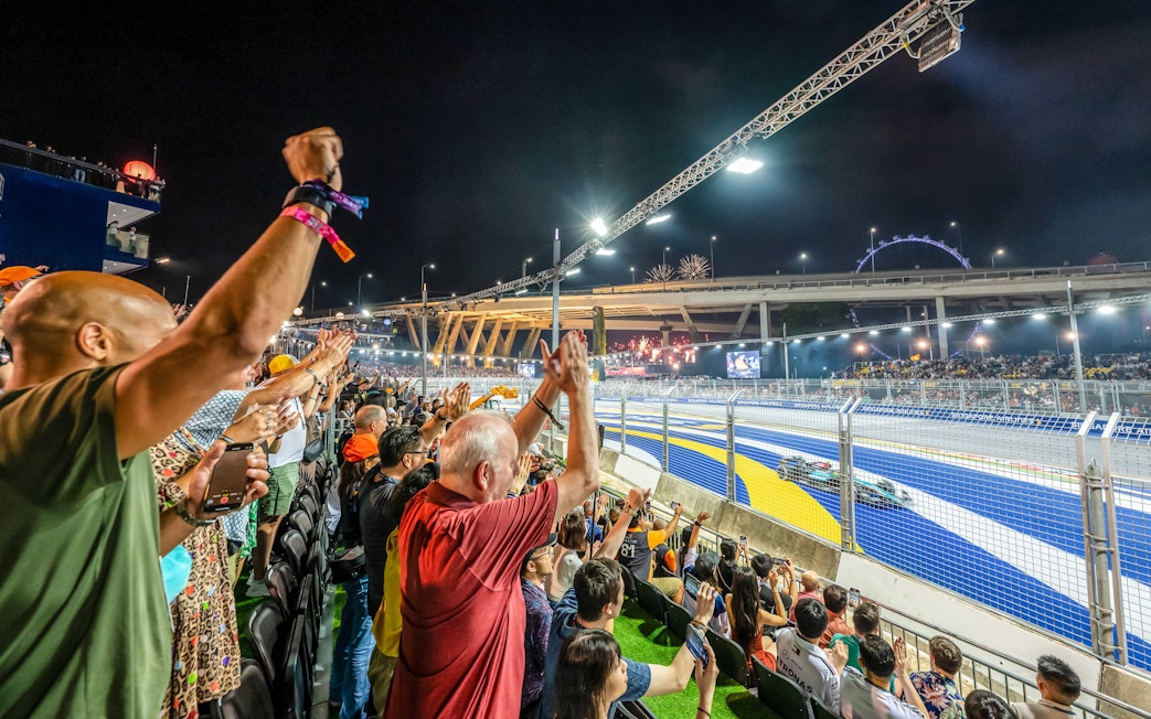 Spectators cheering at a night race with fireworks and city skyline in the background.