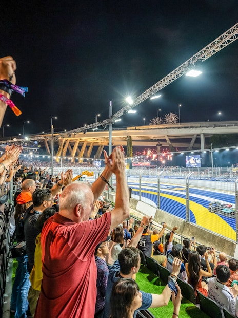 Spectators cheering at a night race with fireworks and city skyline in the background.