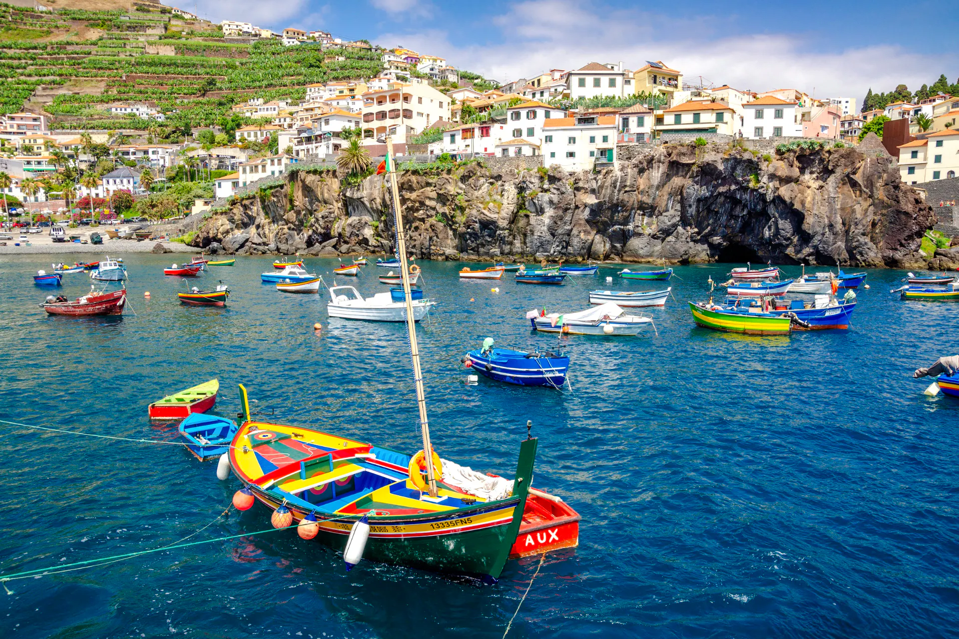 Colorful boats in the harbor of a coastal village in Madeira, with hillside houses in the background.