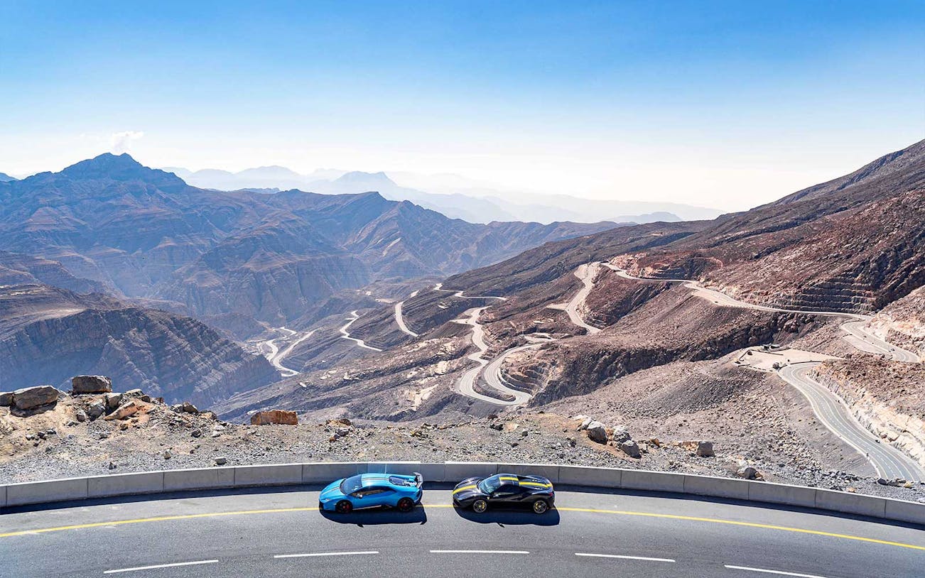 Supercars driving on winding roads at Jabal Bil Ays, surrounded by rugged mountain landscape.