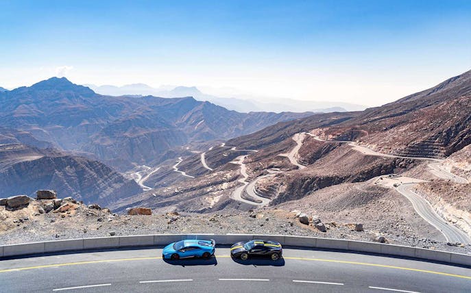 Supercars driving on winding roads at Jabal Bil Ays, surrounded by rugged mountain landscape.
