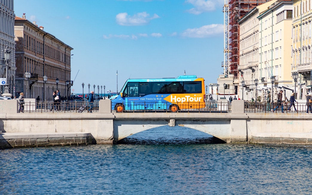 Hop-on hop-off bus crossing bridge in Trieste with historic buildings and Adriatic Sea in view.