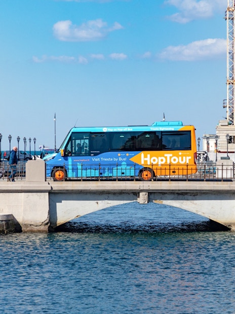 Hop-on hop-off bus crossing bridge in Trieste with historic buildings and Adriatic Sea in view.