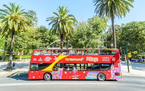 Red double-decker bus for Malaga hop-on hop-off tour near palm trees.