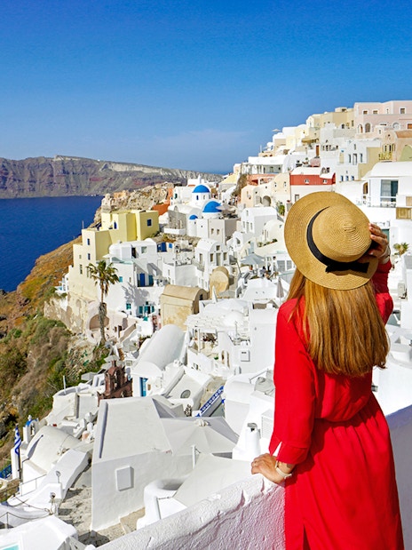 Woman in red dress overlooking white buildings and sea in Oia, Santorini.