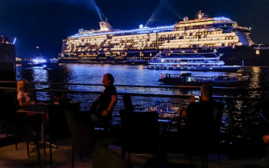Large illuminated cruise ship on evening water, viewed from a waterfront dining area.