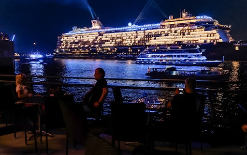 Large illuminated cruise ship on evening water, viewed from a waterfront dining area.