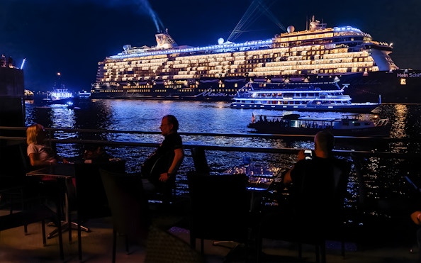 Large illuminated cruise ship on evening water, viewed from a waterfront dining area.