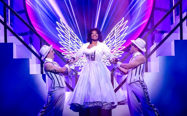 Performer in angel costume with neon wings on stage during Grease musical.