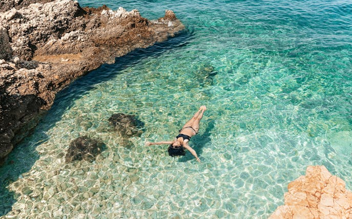 Woman floating in clear turquoise sea near rocky shore in Athens.