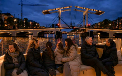 People enjoying a boat tour during the Amsterdam Light Festival with illuminated bridge in view.