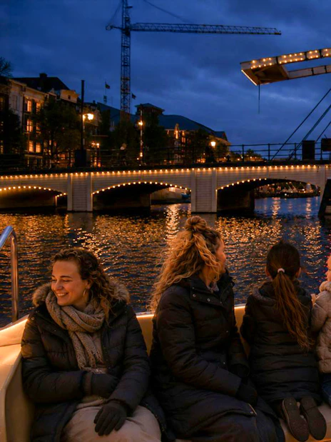 People enjoying a boat tour during the Amsterdam Light Festival with illuminated bridge in view.