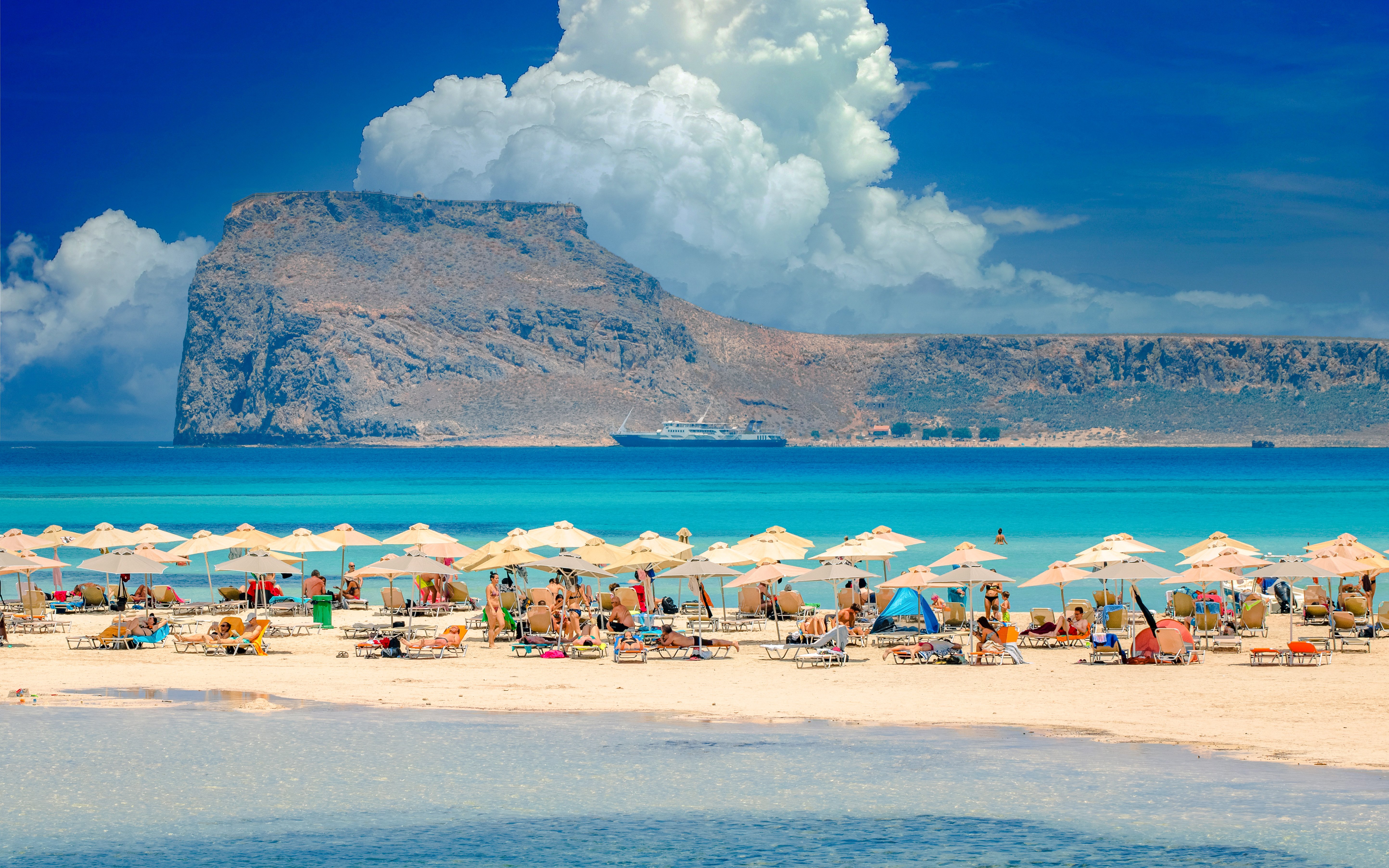 Beachgoers under umbrellas at Balos Lagoon, Crete, with a ship and rocky island in the background.