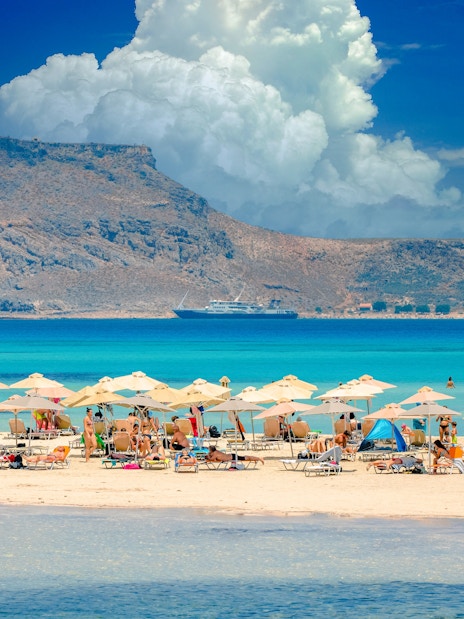 Beachgoers under umbrellas at Balos Lagoon, Crete, with a ship and rocky island in the background.