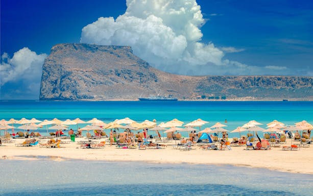 Beachgoers under umbrellas at Balos Lagoon, Crete, with a ship and rocky island in the background.