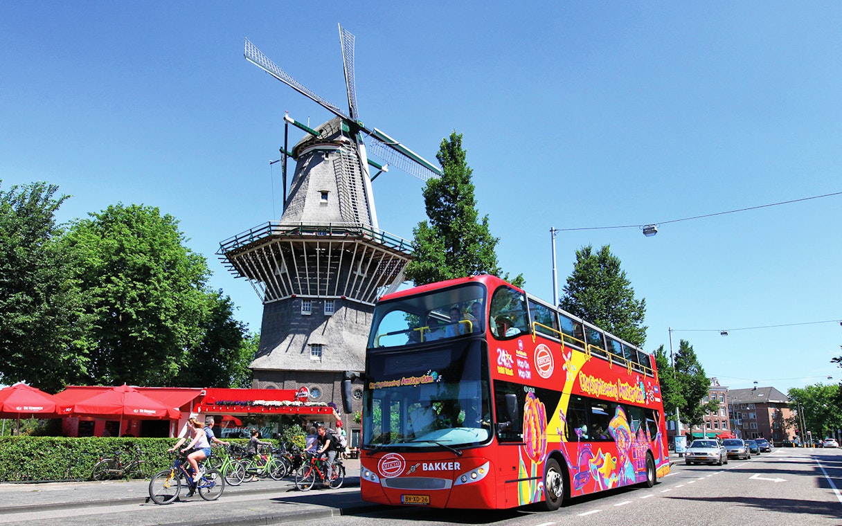 Red double-decker bus on Amsterdam street near a traditional windmill.