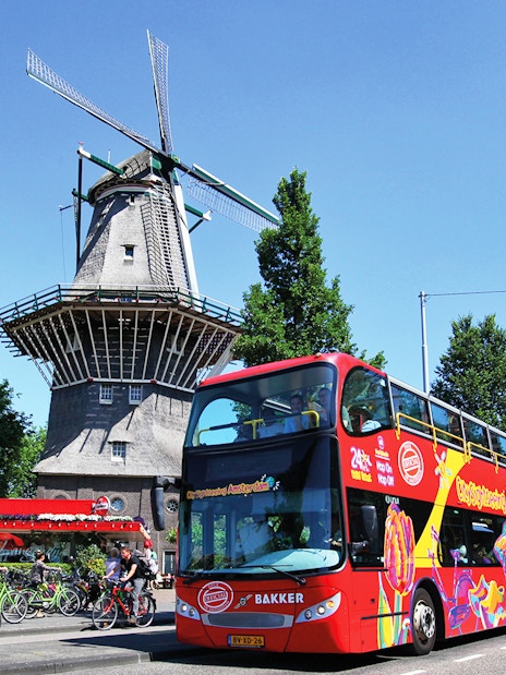 Red double-decker bus on Amsterdam street near a traditional windmill.