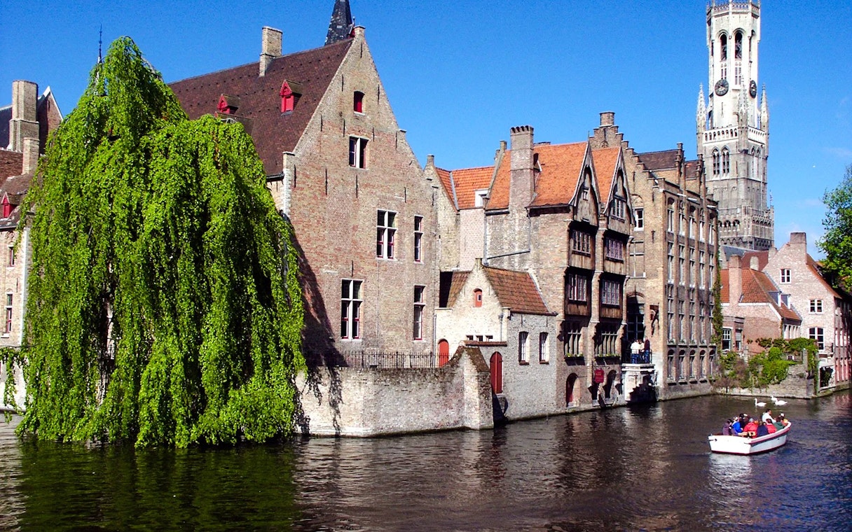 Boat tour on the canals of Brugge with historic buildings and Belfry tower in view.