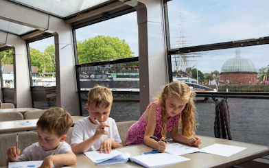 Children drawing on a London city cruise with the Cutty Sark and Greenwich visible outside.