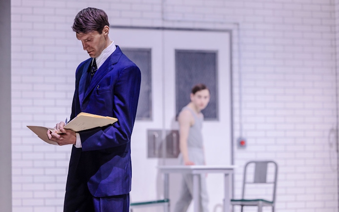Man in blue suit writing on clipboard, another person in background, white brick wall setting.