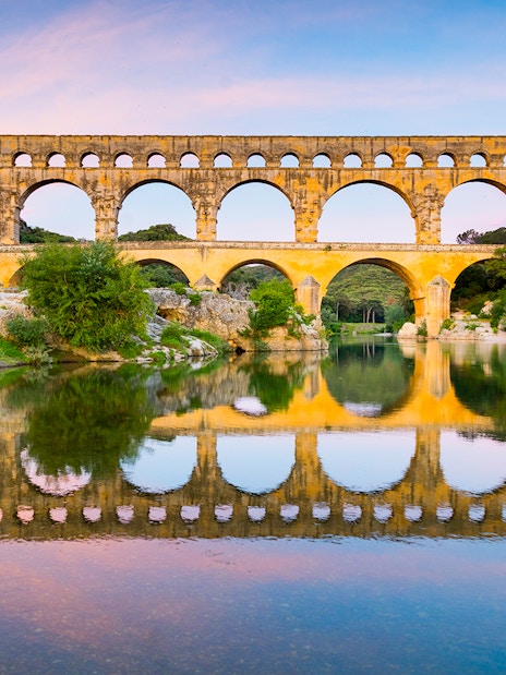Pont du Gard aqueduct reflecting in the Gardon River, France, with surrounding greenery.