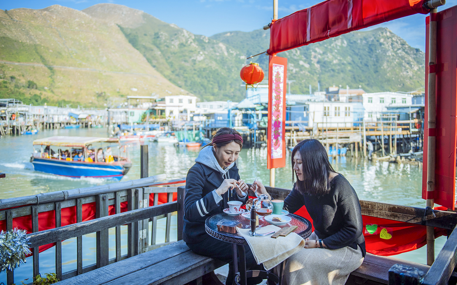 Visitors enjoying tea at a waterfront café on Lantau Island with boats and mountains in the background.