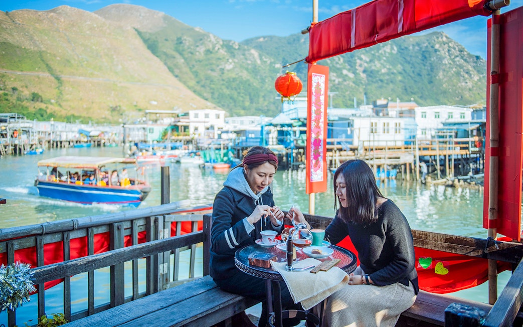 Visitors enjoying tea at a waterfront café on Lantau Island with boats and mountains in the background.