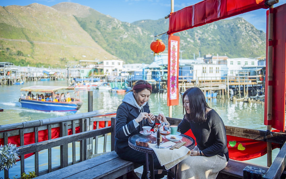 Visitors enjoying tea at a waterfront café on Lantau Island with boats and mountains in the background.
