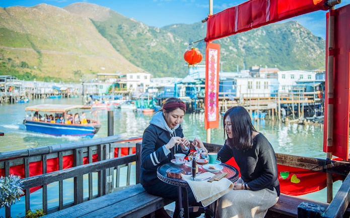 Visitors enjoying tea at a waterfront café on Lantau Island with boats and mountains in the background.