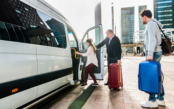 Driver helping woman board van at airport with luggage nearby.