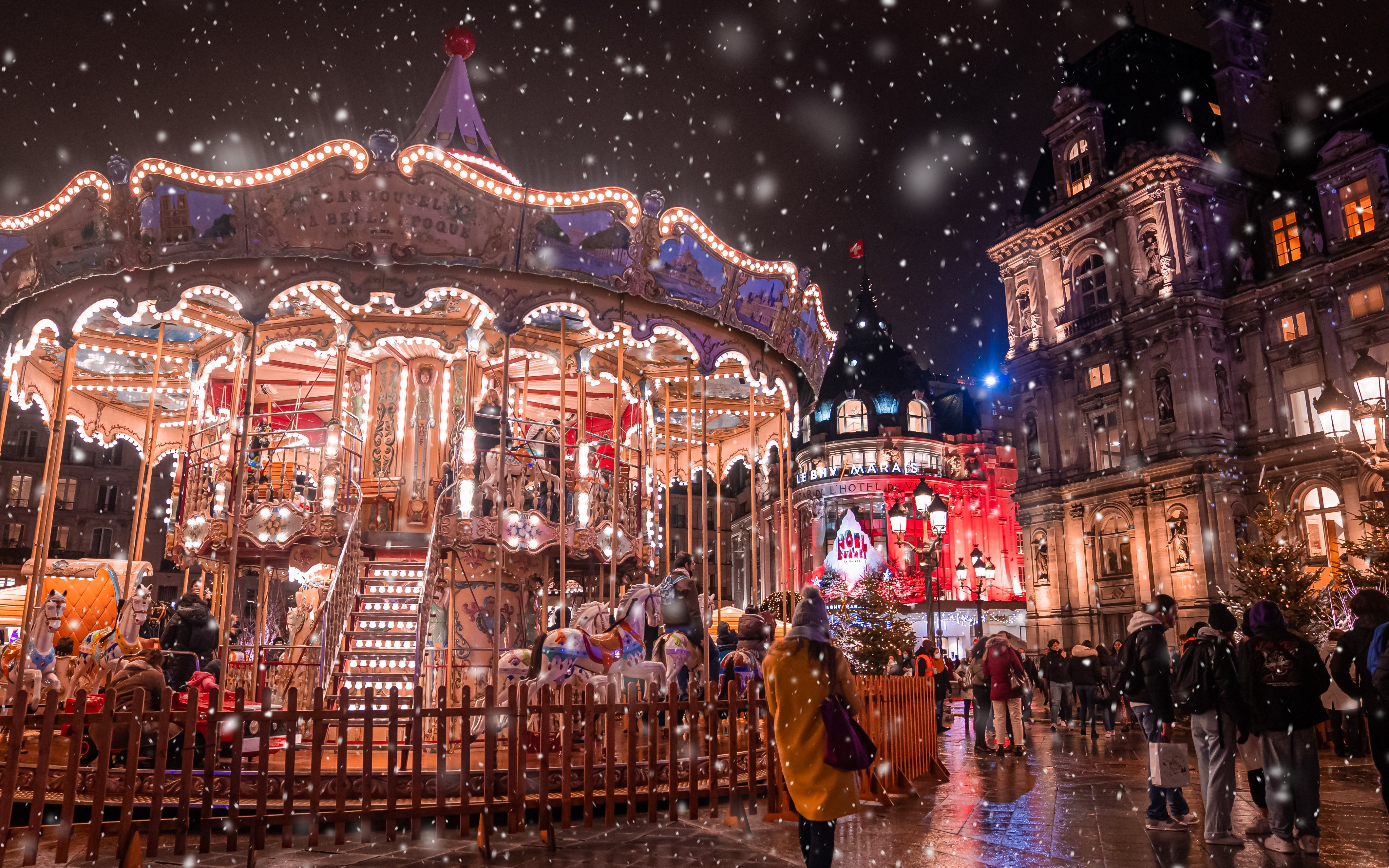 Carousel at night with festive lights and snow in front of Parisian buildings during Christmas.