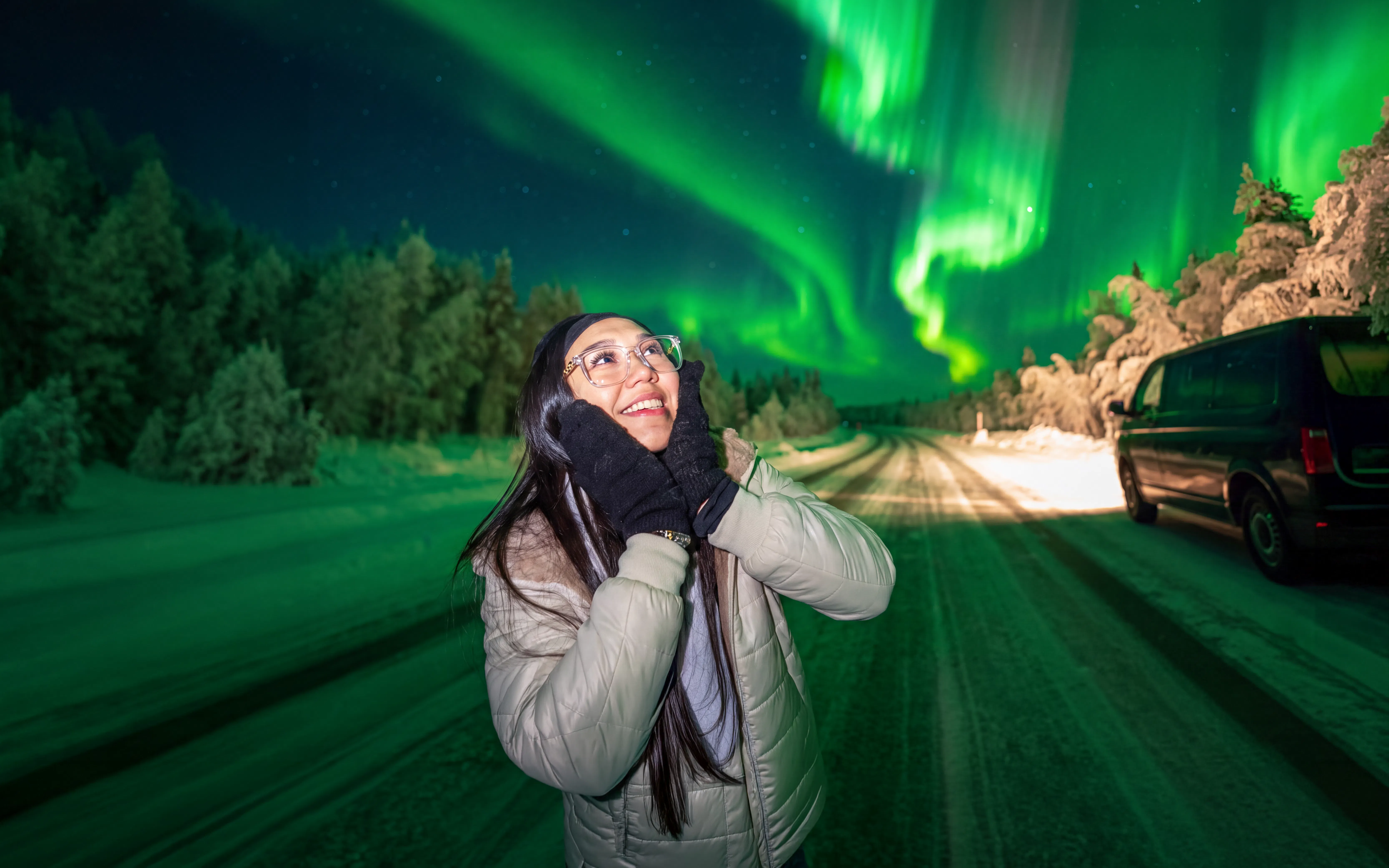 Person enjoying the Northern Lights on a photography tour, snowy road and van in background.