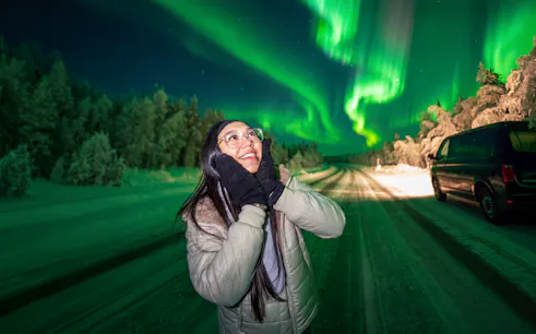 Person enjoying the Northern Lights on a photography tour, snowy road and van in background.
