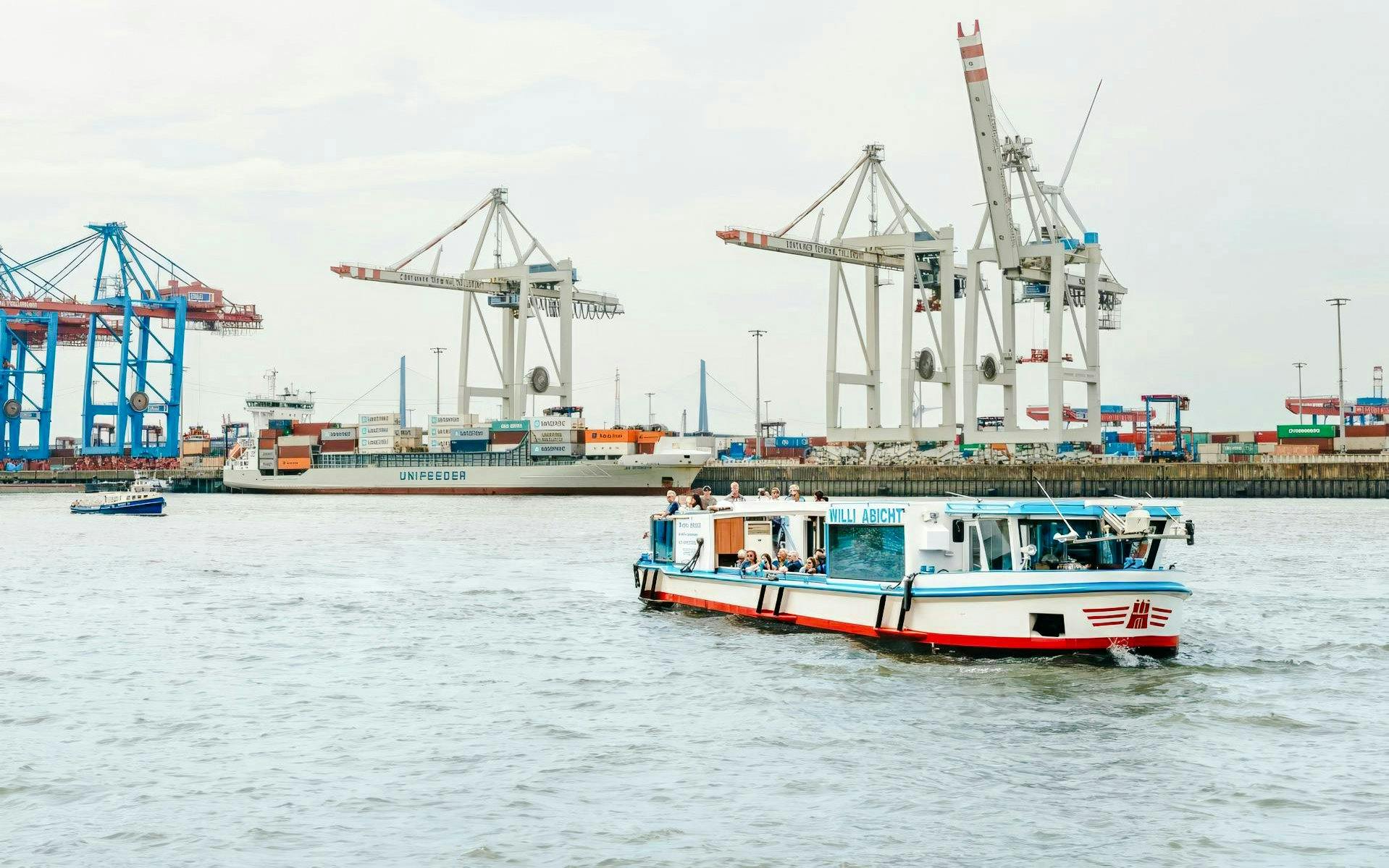 Cruise boat on Hamburg port with cranes and container ship in background.