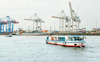Cruise boat on Hamburg port with cranes and container ship in background.