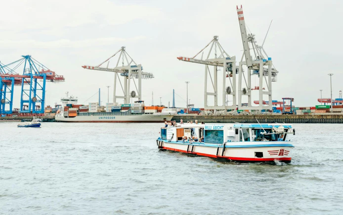 Cruise boat on Hamburg port with cranes and container ship in background.