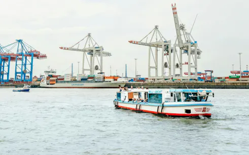 Cruise boat on Hamburg port with cranes and container ship in background.