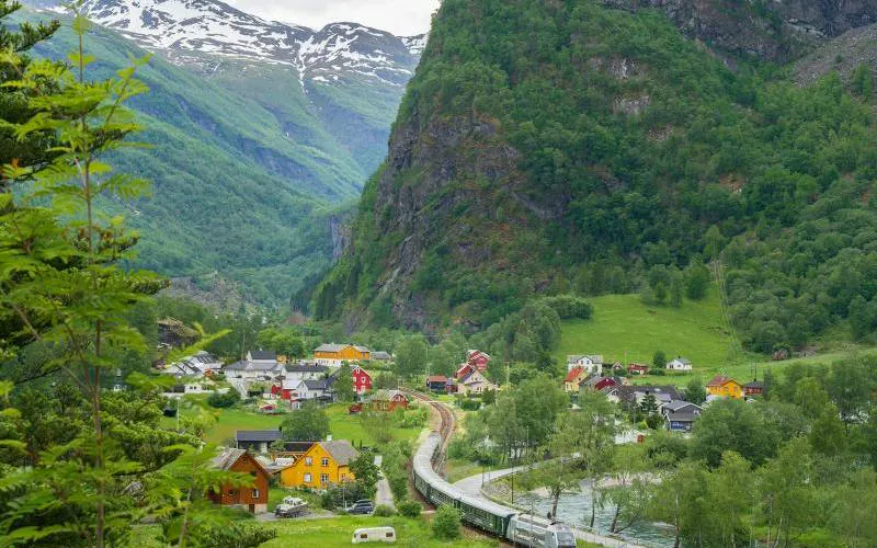 Train traveling through lush valley with colorful houses and mountains, Bergen to Flåm Railway, Norway.