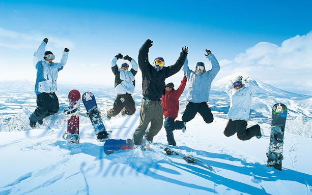 Group of snowboarders jumping on snowy mountain in Rusutsu, Japan, with snowboards in the foreground.