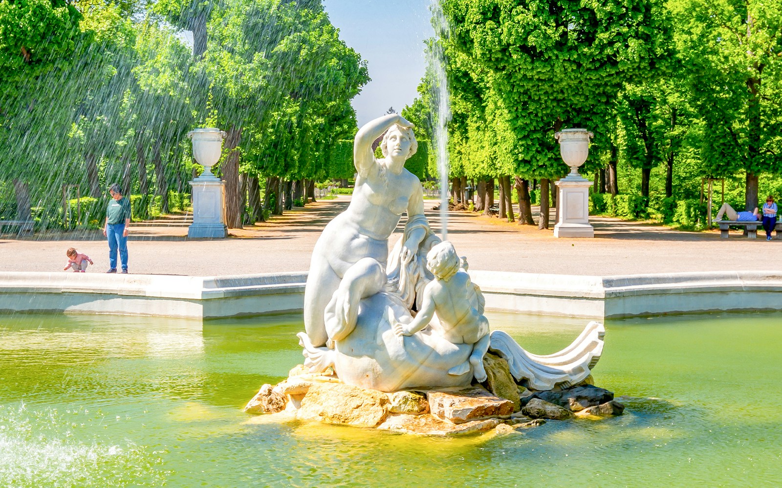 Fountain sculpture in Schönbrunn Palace gardens, Vienna, with lush greenery and visitors nearby.