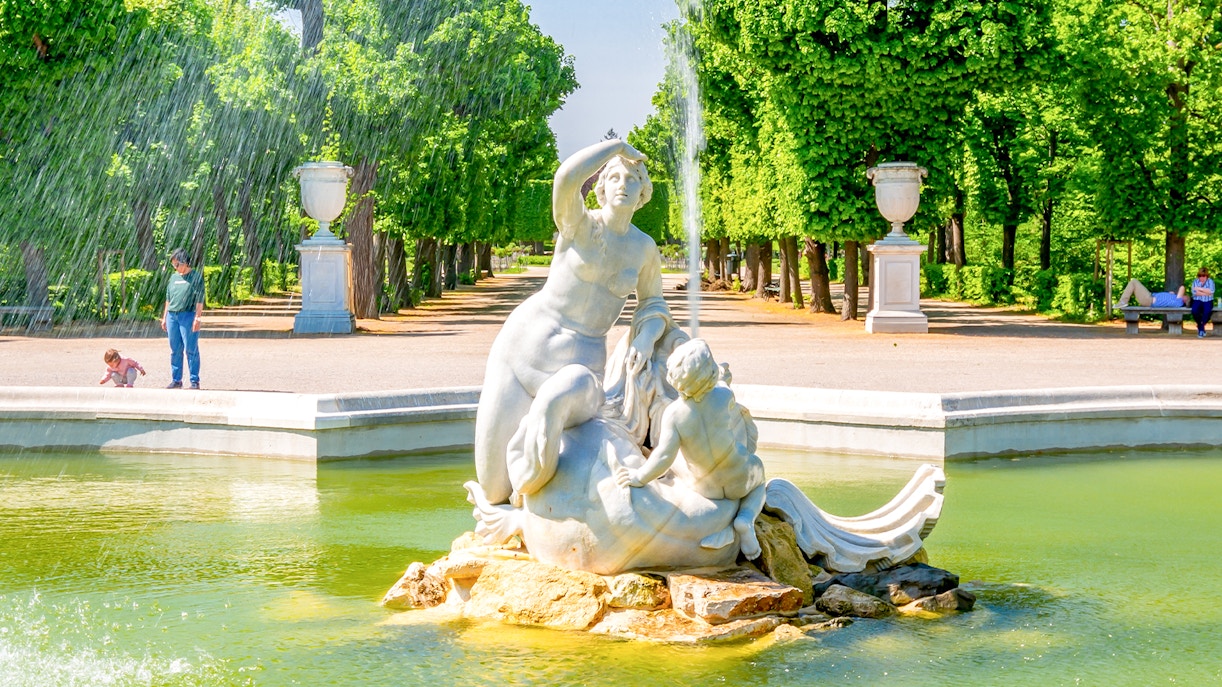 Fountain sculpture in Schönbrunn Palace gardens, Vienna, with lush greenery and visitors nearby.