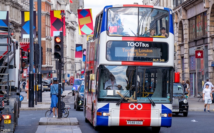 Toot bus navigating a busy street in London with colorful banners overhead.