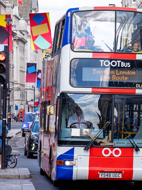 Toot bus navigating a busy street in London with colorful banners overhead.